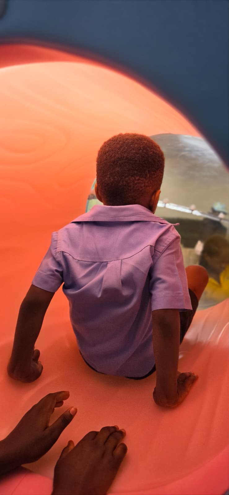 A child seen from behind, sitting inside Letzplay's orange tunnel slide, looking out through its bubble window.